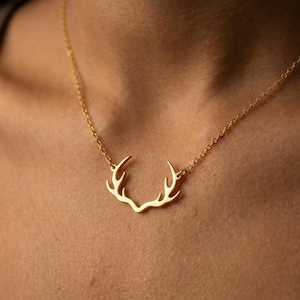 May include: A gold-colored necklace featuring a stylized antler pendant. The pendant is a delicate, openwork design, suspended from a fine chain. The necklace is shown against a skin tone background.