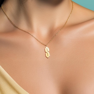 May include: A gold-coloured necklace featuring a pendant in the shape of the letter "S". The pendant has a geometric design. The necklace is worn around the neck.