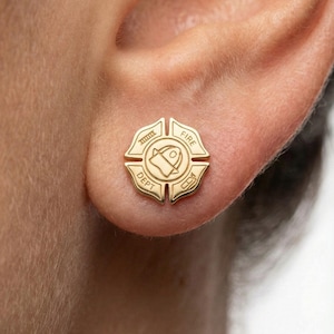 May include: A gold-colored stud earring in the shape of a fire department badge. The earring is worn on an ear. The badge design includes the words "FIRE" and "DEPT" and a central emblem.