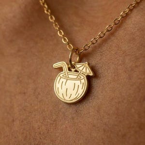 May include: A gold necklace with a pendant depicting a coconut drink with a straw and a small umbrella. The gold chain is visible against a skin-toned background. The pendant is a tropical-themed charm.
