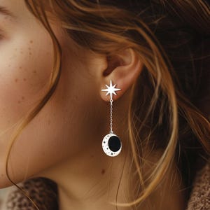 Puede incluir: Pendientes plateados con temática celestial. Cada pendiente presenta una estrella, una fina cadena y un colgante de media luna con centro negro y detalles de estrellas. Los pendientes están diseñados para colgar, añadiendo un toque de fantasía.