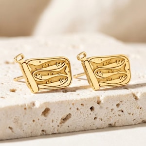 May include: A pair of gold-toned stud earrings shaped like rectangular sardine tins. Each earring features an engraved design of three small fish stacked horizontally inside the tin, complete with a small pull-tab detail at the top corner.