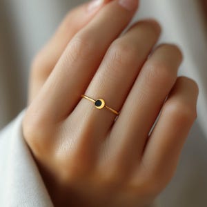 Puede incluir: Un delicado anillo de oro con un pequeño diseño circular negro. El anillo es fino y minimalista, con un acabado pulido. El anillo se muestra en un dedo, destacando su simple elegancia. Una joya.