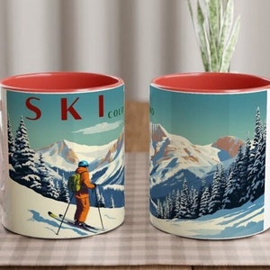 Puede incluir: Dos tazas de cerámica con interior rojo y una escena de esquí invernal. Las tazas presentan un esquiador, montañas cubiertas de nieve y la palabra "SKI COLORADO" en rojo. El fondo es un cielo azul con nubes blancas.