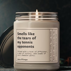 May include: A 9 ounce soy wax candle with a white label that reads "Smells like the tears of my tennis opponents. Infused with a hint of A**kicker, a dash of victory, and loads of court champion glory." The candle is in a clear glass jar with a black lid.