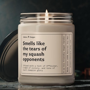 May include: A 9 ounce soy wax candle in a glass jar with a beige label. The label reads "Smells like the tears of my squash opponents" and "Infused with a hint of A**kicker, a dash of victory, and tons of court champion glory". The candle is made with 100% natural soy wax and is eco-friendly and vegan.