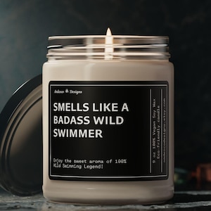May include: A white candle in a glass jar with a black label that reads "Smells Like a Badass Wild Swimmer" and "Enjoy the sweet aroma of 100% Wild Swimming Legend!" The candle is lit and the flame is visible.
