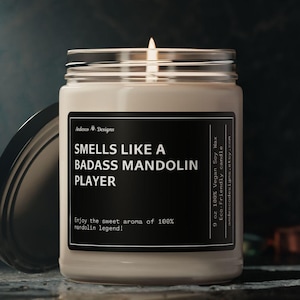 May include: A white candle in a glass jar with a black label that reads "Smells Like A Badass Mandolin Player" and "Enjoy the sweet aroma of 100% mandolin legend!" The candle is 9 oz and made with 100% vegan soy wax.