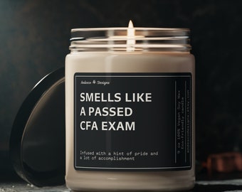 May include: A beige soy candle in a glass jar with a black label that reads "Smells Like A Passed CFA Exam". The candle is lit and the flame is visible. The text on the label also reads "Infused with a hint of pride and a lot of accomplishment".