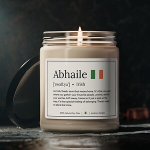 Może przedstawiać: Beżowa świeca w szklanym słoiku z białą etykietą. Etykieta zawiera tekst "Abhaile ['awall:ya']  Irish" i małą irlandzką flagę. Tekst na etykiecie brzmi: "Irlandzkie słowo gaelickie oznaczające dom. To przytulne miejsce, w którym gromadzisz swoich ulubionych ludzi, odpoczywasz i pozwalasz, by twoje troski zniknęły. Dom to nie tylko punkt na mapie, to szczególne uczucie przynależności. Nie ma miejsca takiego jak dom. 100% naturalny wosk sojowy | Andesco Designs"