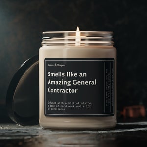 May include: A beige candle in a glass jar with a black label that reads "Smells like an Amazing General Contractor. Infused with a hint of vision, a dash of hard work and a lot of excellence." The candle is lit and the flame is visible.