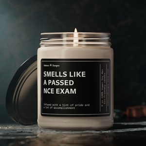 May include: A white candle with a black label that reads "Smells Like A Passed NCE Exam" and "Infused with a hint of pride and a lot of accomplishment". The candle is in a glass jar with a black lid.
