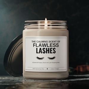 Może przedstawiać: Beżowa świeca z czarną etykietą, na której widnieje napis "THE CALMING SCENT OF FLAWLESS LASHES" wraz z grafiką rzęs. Etykieta zawiera również napis "Infused with style, charm and jaw-dropping looks" oraz "100% Natural Soy Wax | Andesco Designs".