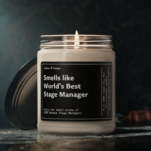 May include: A white candle with a black label that reads "Smells like World's Best Stage Manager!" Enjoy the sweet aroma of 100% Badass Stage Manager!"