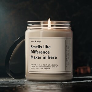 May include: A beige candle with a glass jar and a white label that reads "Smells like Difference Maker in here". The label also includes the text "Infused with a hint of vision, a dash of determination and a lot of positive impact".