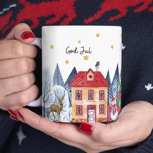 Puede incluir: Taza de cerámica blanca con una ilustración de escena de invierno. La taza presenta una casa con techo rojo, un ciervo, un muñeco de nieve y un pájaro. El texto "God Jul" está escrito en la taza.
