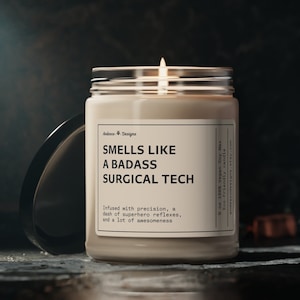 Puede incluir: Una vela de cera de soja blanca en un tarro de vidrio transparente con tapa negra. La vela tiene una etiqueta que dice "Smells Like A Badass Surgical Tech" y "Infused with precision, a dash of superhero reflexes, and a lot of awesomeness".