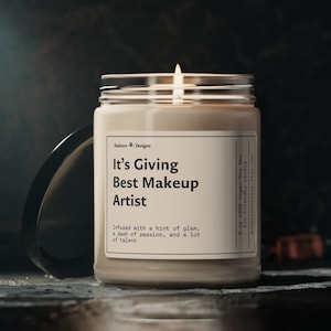 May include: A beige candle in a glass jar with a black lid. The candle is labelled "It's Giving Best Makeup Artist" and "Infused with a hint of glam, a dash of passion, and a lot of talent". The candle is 9 oz and made with 100% vegan soy wax.