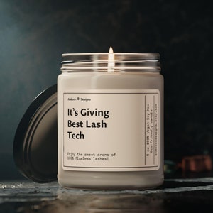 May include: A white candle with a black label that reads "It's Giving Best Lash Tech" and "Enjoy the sweet aroma of 100% flawless lashes!" The candle is in a glass jar with a black lid. The candle is lit and the flame is visible.