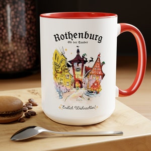 May include: White ceramic mug with a red rim and handle. The mug features a colourful illustration of a German town with the text "Rothenburg Ob der Tauber" and "Endlich Weihnachten!"