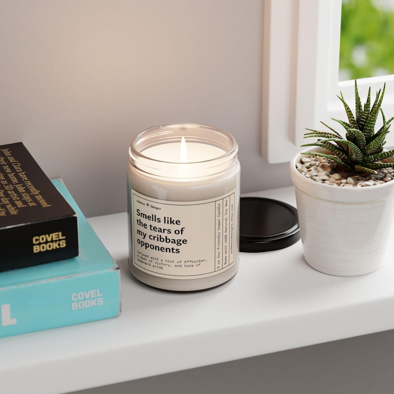 May include: A white candle with a black label that reads "Smells like the tears of my cribbage opponents". The candle is lit and sitting on a shelf with two books.