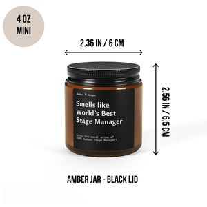 May include: A 4 ounce amber glass candle jar with a black lid. The candle is labelled "Smells like World's Best Stage Manager" and has the text "Enjoy the sweet aroma of 100% Badass Stage Manager!" The jar is 6 cm wide and 6.5 cm tall.