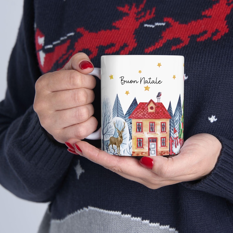 May include: White ceramic mug with a winter scene and the text "Buon Natale" in black. The scene features a yellow house with a red roof, a deer, a snowman, and trees.