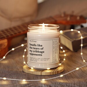 May include: A 9 ounce soy wax candle in a clear glass jar with a white label that reads "Smells like the tears of my cribbage opponents. Infused with a hint of A**kicker, a dash of victory, and tons of pegboard pride." The candle is lit and sitting on a wooden coaster.