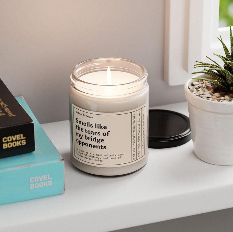 May include: A white candle with a black lid and a label that reads "Smells like the tears of my bridge opponents. Infused with a hint of A**kicker, a dash of victory, and a tone of bridge master pride."