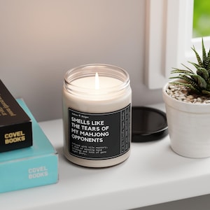 May include: A white candle with a black label that reads "Smells like the tears of my Mahjong opponents." The label also includes the text "Infused with tile mastery, a dash of winning hand strategy, and lots of game night glory."
