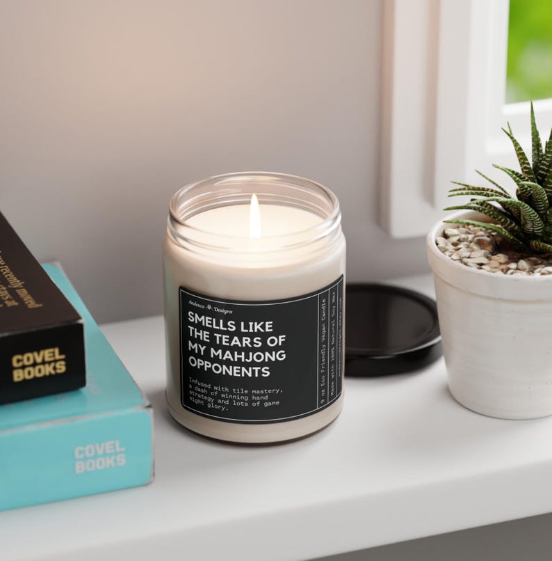 May include: A white candle with a black label that reads "Smells like the tears of my Mahjong opponents." The label also includes the text "Infused with tile mastery, a dash of winning hand strategy, and lots of game night glory."