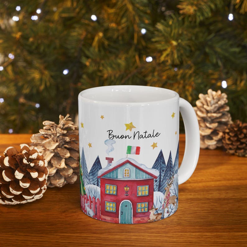 May include: White ceramic mug with a winter scene featuring a red house with a green roof and snow on the roof. The house has a chimney with smoke coming out of it. There are trees in the background and the text "Buon Natale" is written on the mug.