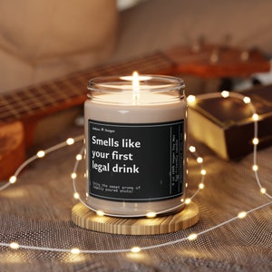 May include: A brown soy candle in a clear glass jar with a black label that reads "Smells like your first legal drink" and "Enjoy the sweet aroma of legally poured shots!" The candle is on a wooden coaster and surrounded by white string lights.