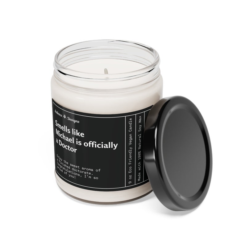 K&ouml;nnte beinhalten: Eine 9 Unzen Sojawachskerze in einem durchsichtigen Glasgef&auml;&szlig; mit schwarzem Deckel. Das Etikett lautet "Smells like Michael is officially a Doctor" und "Eco Friendly Vegan Candle".