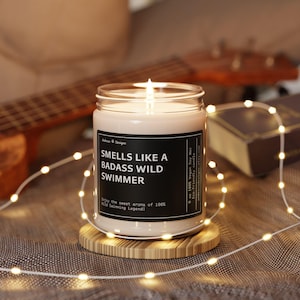 May include: A white soy wax candle in a clear glass jar with a black label that reads "Smells Like A Badass Wild Swimmer". The candle is lit and sitting on a wooden coaster.