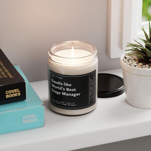 May include: A clear glass candle with a white wax base and a black label that reads "Smells like World's Best Stage Manager". The candle is lit and the flame is visible. The candle is sitting on a white surface.