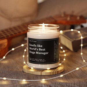 May include: A white soy wax candle in a clear glass jar with a black label that reads "Smells like World's Best Stage Manager". The candle is lit and sitting on a wooden coaster.