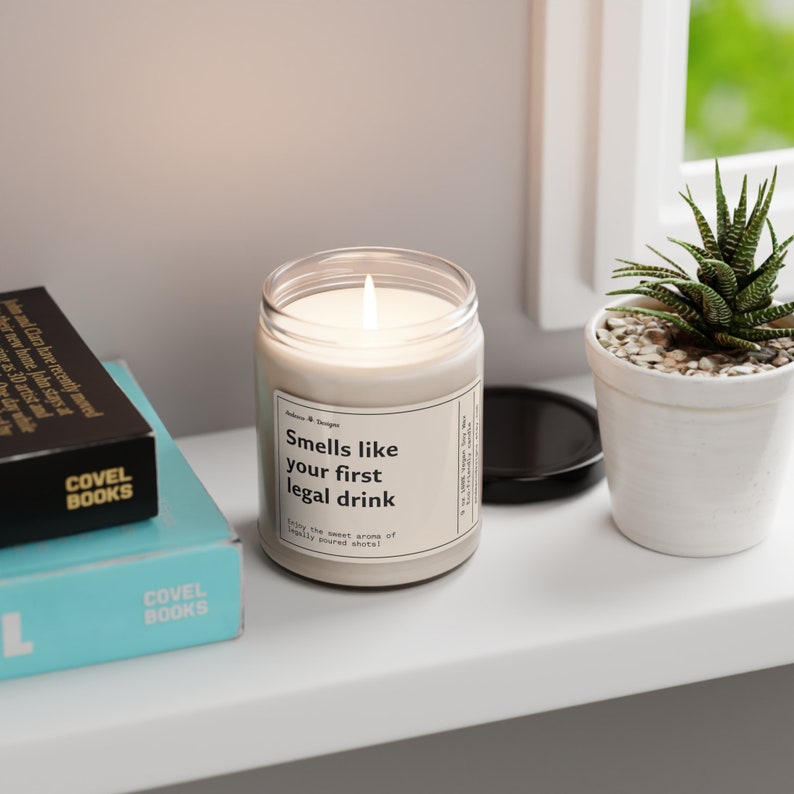 May include: A white candle with a black label that reads "Smells like your first legal drink. Enjoy the sweet aroma of legally poured shots!" The candle is on a white shelf with a book and a potted plant.