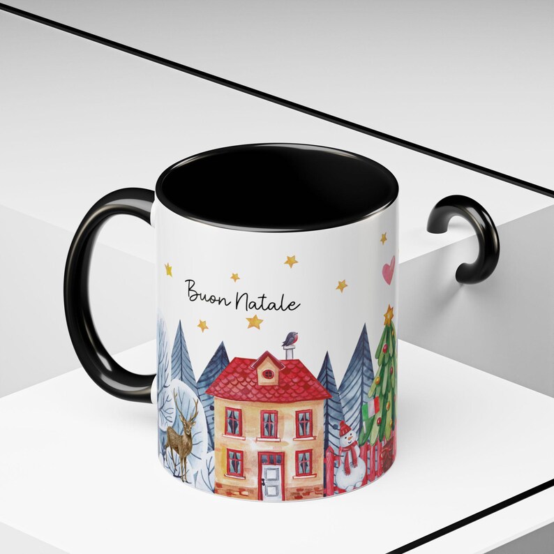 May include: A white ceramic mug with a black handle and rim. The mug features a winter scene with a house, trees, a snowman, a deer, and a bird. The text "Buon Natale" is written in black on the mug.