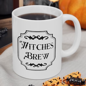 May include: White ceramic mug with a black and white graphic that says "Witches Brew". The mug is filled with coffee and there are orange and black candies scattered around it.