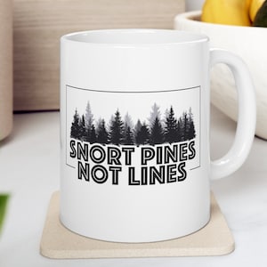 May include: White ceramic mug with a black and gray silhouette of a pine forest and the text "Snort Pines Not Lines".