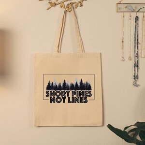 May include: A beige tote bag with a black and white graphic of pine trees and the text "Snort Pines Not Lines".