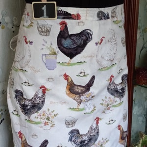 May include: A white apron with a black ruffle trim and a chicken print. The apron has a pocket with a small chalkboard sign that says "1". The print features various breeds of chickens, including Australorp, White Frizzle, and Brown Leghorn.