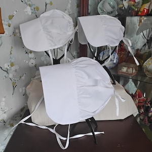 May include: Three white bonnets with ruffled edges and ties. The bonnets are displayed on a wooden table.
