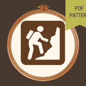 May include: Cross-stitch pattern featuring a hiker climbing a mountain, displayed in a wooden embroidery hoop. The design is in brown and white, with the text "PDF PATTERN" on a green banner.