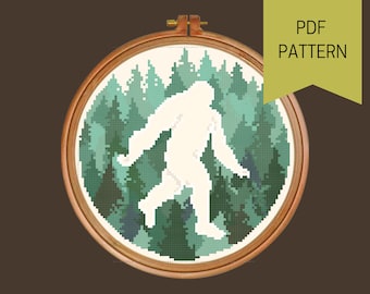 May include: A cross-stitch pattern featuring a white silhouette of Bigfoot walking through a forest of green trees. The pattern is in a wooden embroidery hoop.