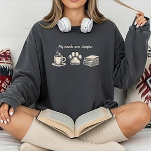 May include: Dark gray sweatshirt with the phrase "My needs are simple" and illustrations of a coffee cup, paw print, and books. The sweatshirt is worn with headphones and a book.