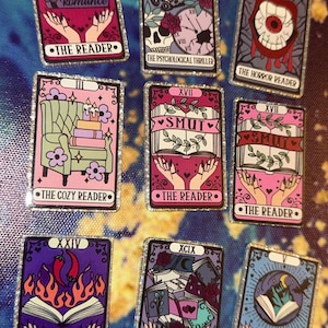 May include: A collection of nine rectangular enamel pins designed like tarot cards, featuring various illustrations including skulls, books, eyes, and candles. Each pin has a silver glitter border and text labels such as The Reader, Smut, and The Collector.