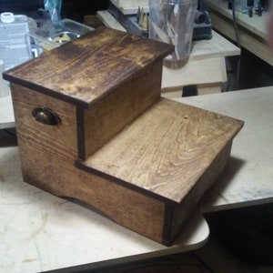 Puede incluir: Un taburete de madera de dos peldaños con un tinte marrón oscuro. El taburete tiene un pequeño cajón con una perilla de latón en la parte delantera. El taburete está sentado sobre una superficie de madera clara.