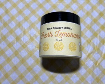 Può includere: Un barattolo di slime da 170 ml etichettato "High Quality Slimes Fresh Lemonade" con tre fette di limone giallo e un bicchiere di limonata sull'etichetta.